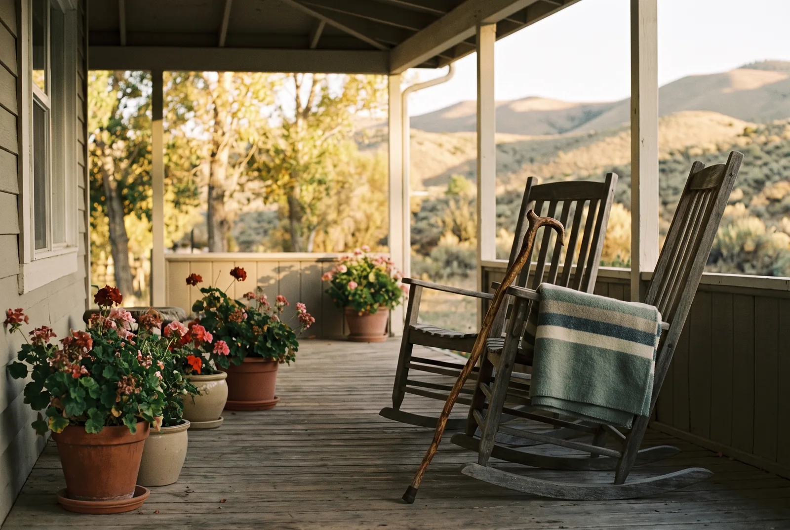 Two empty rocking chairs on a covered porch at golden hour, a cane and a folded blanket resting on them, geraniums alongside